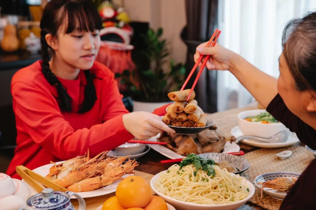 A young woman in a red sweater watches as another person uses bright red chopsticks to lift a golden-brown spring roll from a stacked plate. The table is filled with a festive Lunar New Year feast, including large prawns, noodles, citrus fruits, and various traditional dishes.