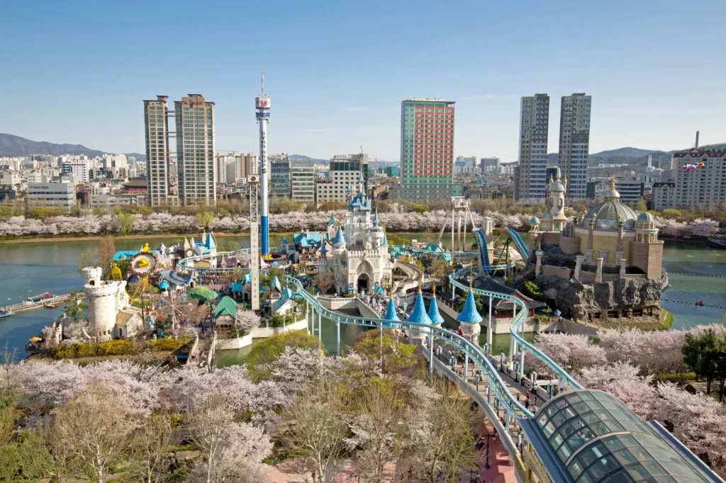 This image captures a vibrant amusement park, likely Lotte World's Magic Island, featuring a fairytale castle and thrilling rides situated on an island in the middle of a lake. The scene is framed by a dense ring of blooming cherry blossoms and a modern city skyline under a clear blue sky.