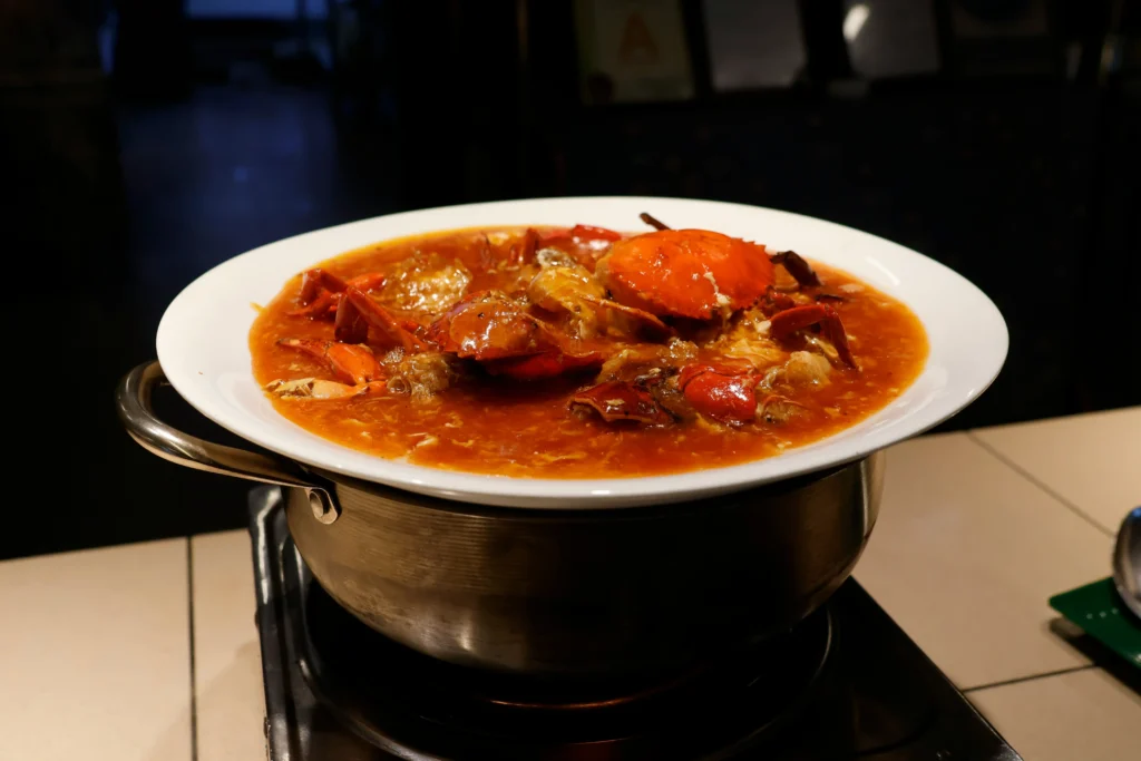 A large, vibrant red crab is served in a rich, savory gravy inside a white bowl. The dish sits atop a metal pot on a portable stove, ready to be enjoyed in a dimly lit setting.