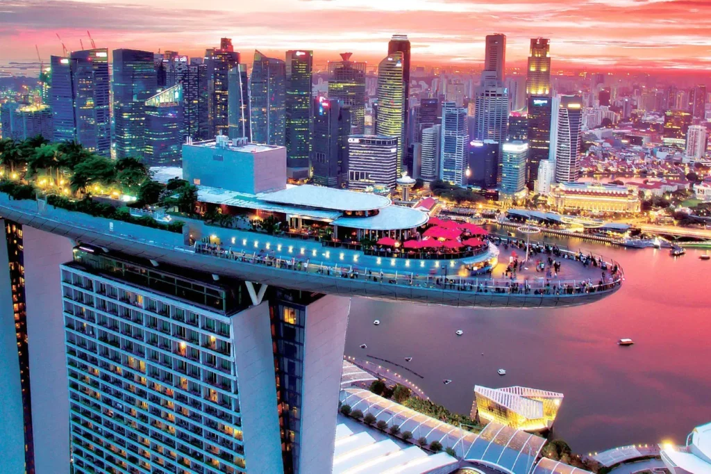 The image showcases a high-angle, sunset view of the Marina Bay Sands SkyPark cantilevered over the Singapore skyline. Crowds of people gather on the observation deck and around red-umbrellaed dining areas, set against a backdrop of illuminated skyscrapers and glowing pink water.