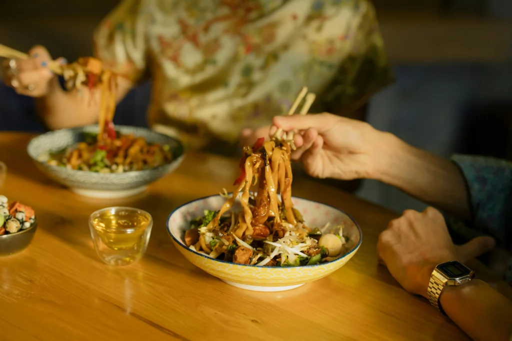 Two people are enjoying bowls of thick, saucy noodles topped with fresh vegetables and sprouts at a wooden table. Using chopsticks, they lift the noodles from decorative ceramic bowls, accompanied by a small glass of tea and light snacks.