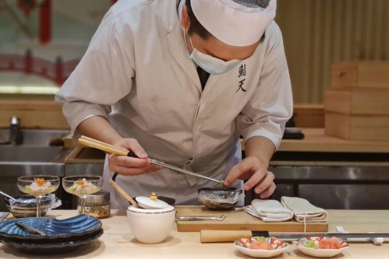A chef wearing a white uniform and a face mask meticulously plates a dish using long metal chopsticks at a wooden sushi counter. The scene features various culinary tools and small bowls of ingredients, highlighting the precision of Japanese fine dining.