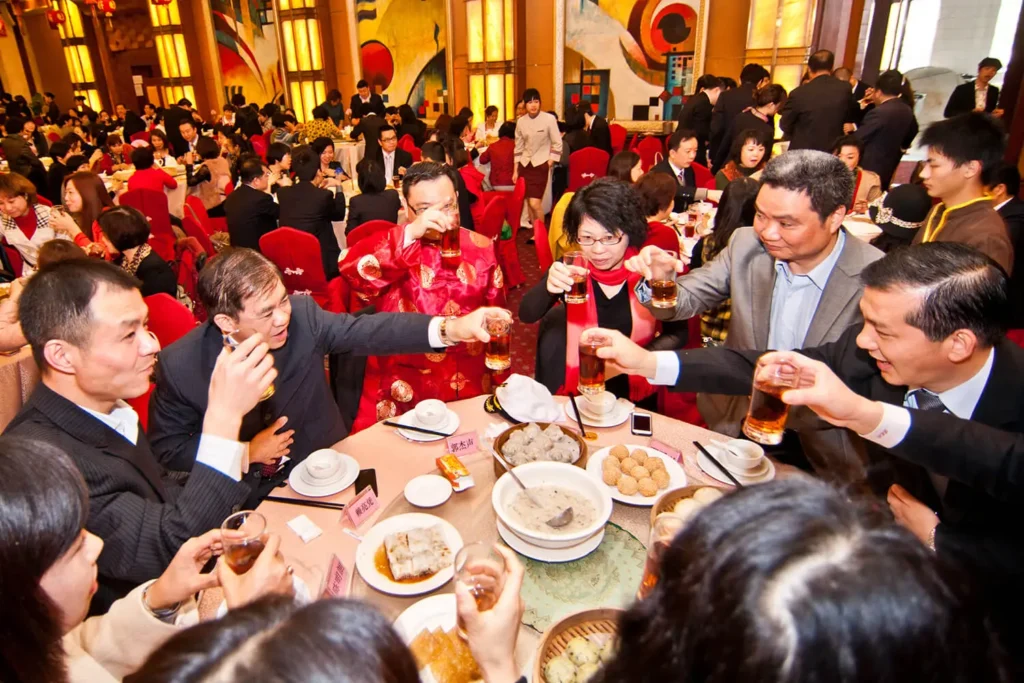 A group of people are gathered around a circular banquet table, raising their glasses in a celebratory toast during a dim sum meal. The crowded restaurant is filled with many other diners and decorated with warm lighting and vibrant red accents.