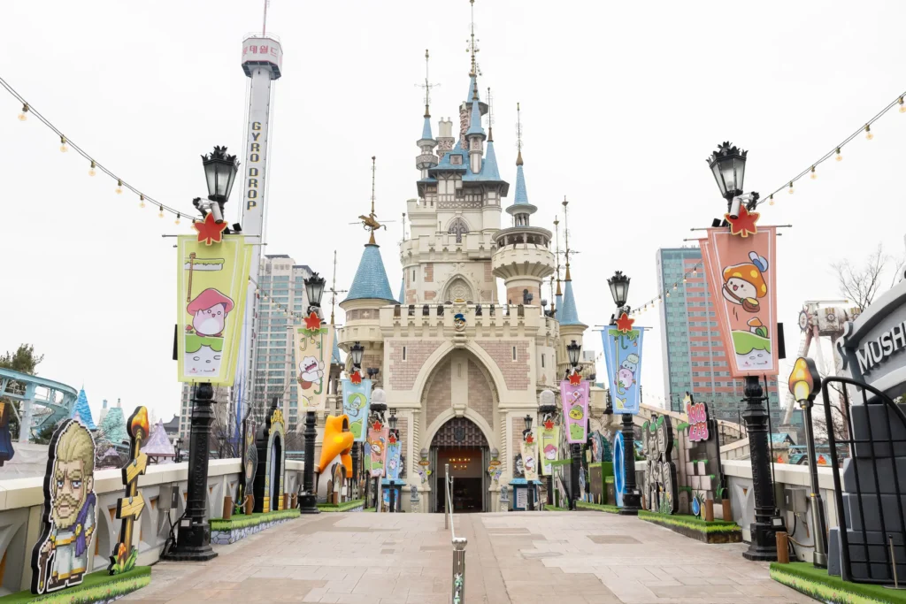 This image shows the fairytale-style Magic Castle at Lotte World in Seoul, decorated with colorful banners and pixel-art characters from the game MapleStory. A paved walkway leads toward the ornate structure, flanked by themed cutouts and a tall "Gyro Drop" tower in the background.