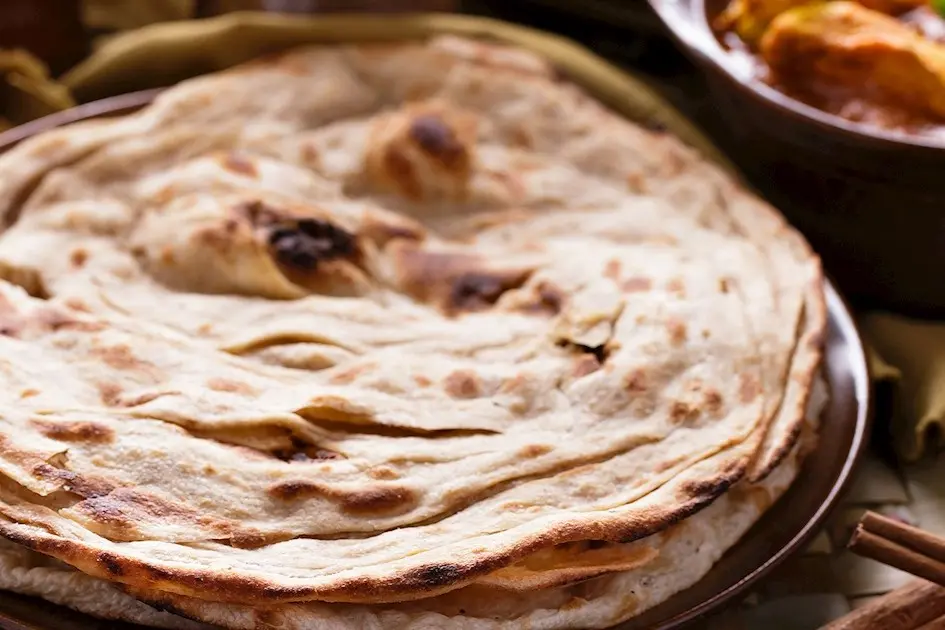 A stack of warm, golden-brown lachha paratha sits on a dark plate, showcasing its characteristic flaky and layered texture. In the background, a bowl of rich curry and cinnamon sticks hint at a flavorful Indian meal.