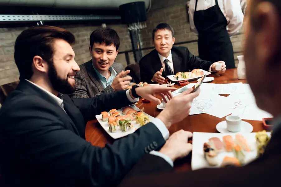 A group of businessmen is gathered around a table in a restaurant, sharing plates of sushi while discussing documents and looking at a smartphone. The atmosphere appears collaborative and professional as they engage in a working lunch or business meeting.