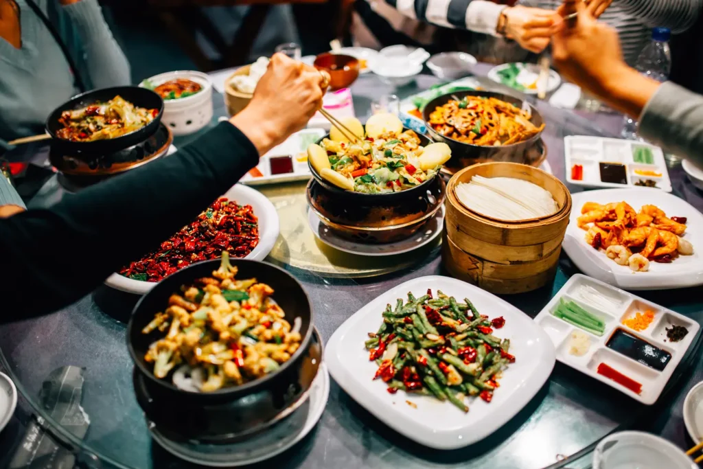 A group of diners uses chopsticks to share a variety of traditional Chinese dishes arranged on a large circular table. The spread includes vibrant stir-fries, steamed buns in bamboo baskets, and assorted condiments, capturing a lively communal dining experience.