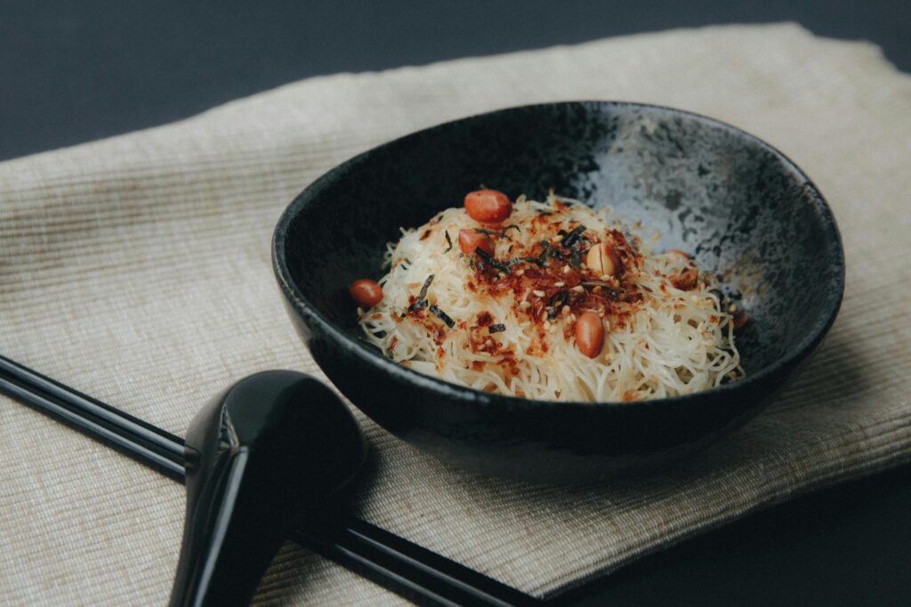 A bowl of thin, chilled vermicelli noodles is topped with whole peanuts, sesame seeds, and dried seaweed. The dish is presented in a dark, textured ceramic bowl resting on a neutral-toned linen cloth alongside a black spoon.