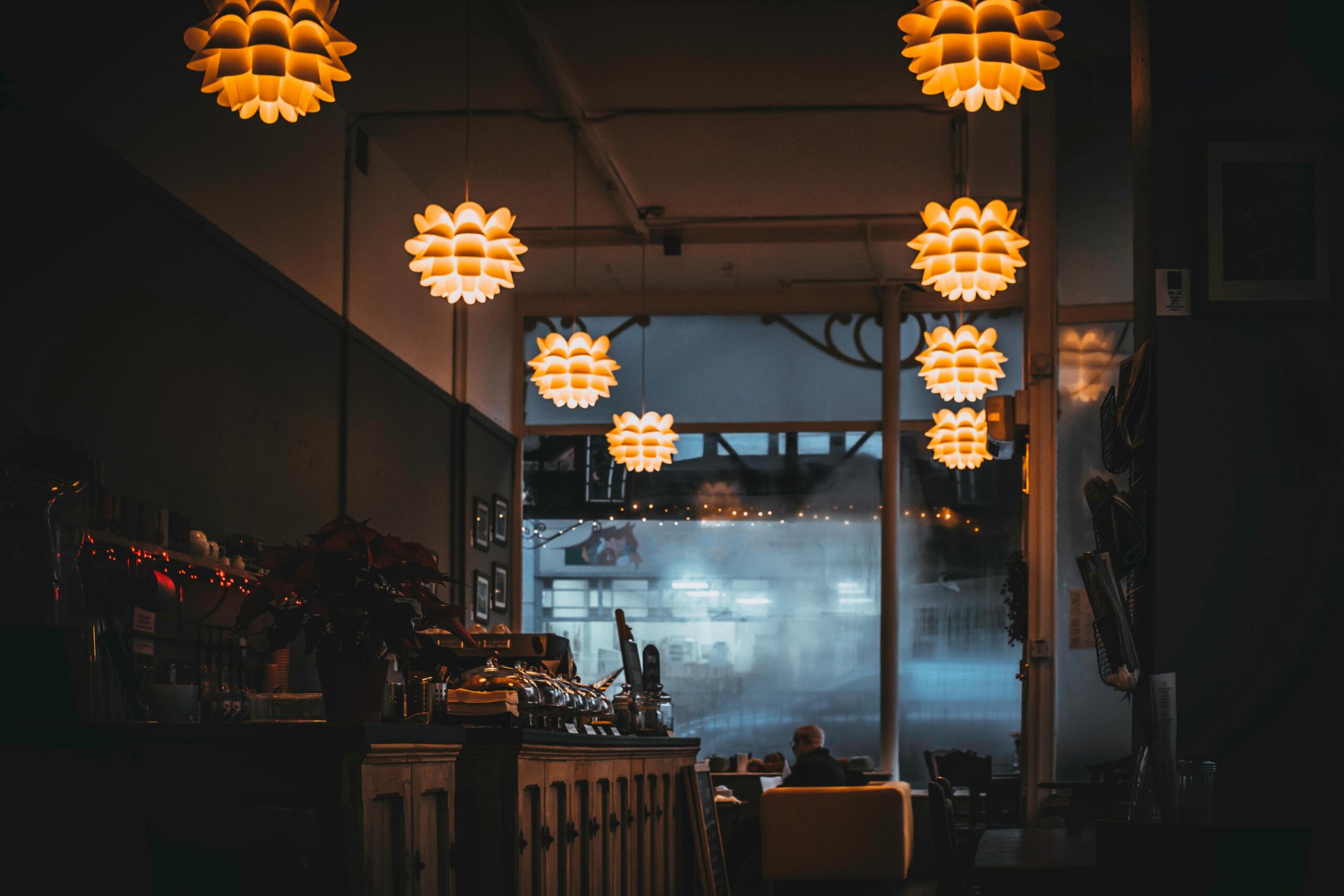 Warm, artichoke-style pendant lights glow in the dim interior of a cozy cafe or restaurant. A person sits at a table in the background, framed by a large window looking out onto a cool, misty street.