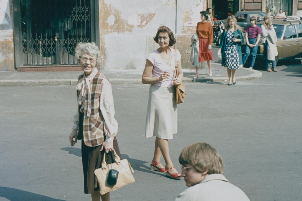 Several women in vintage attire walk across a paved city street, capturing a candid moment from a past era. The scene is filled with retro details, from the classic cars parked in the background to the distinctive patterns and styles of the pedestrians' clothing.