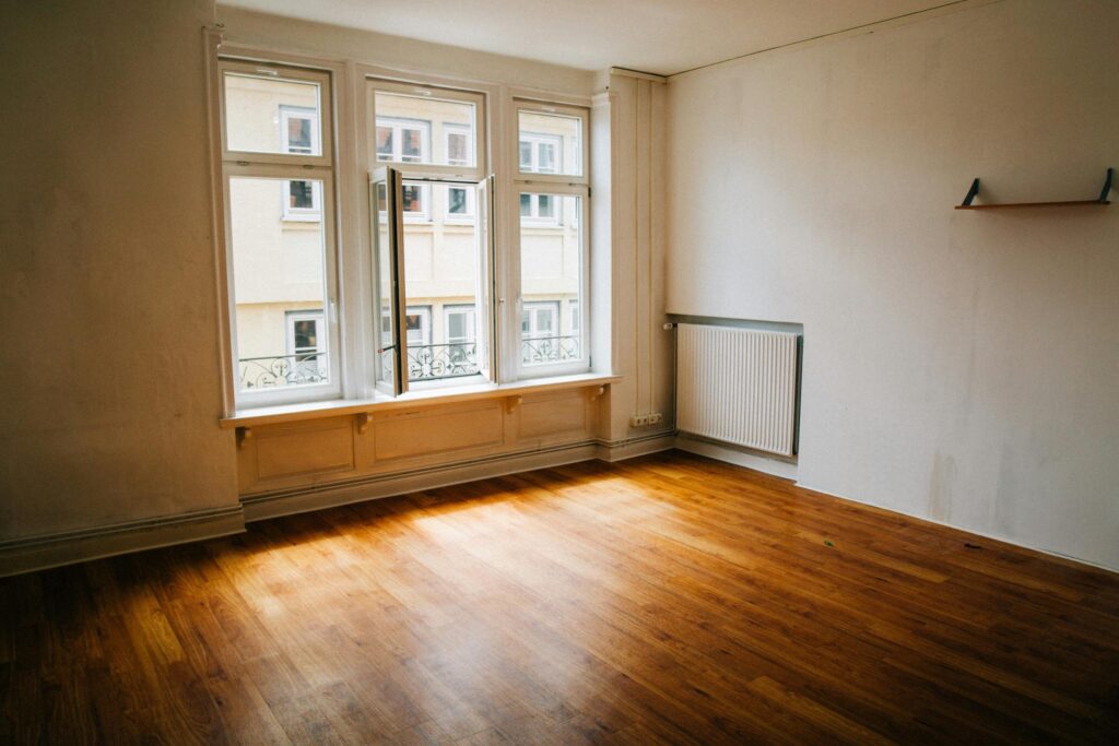 An empty room features warm hardwood flooring and white walls illuminated by natural light from a large triple window. One of the window panes is swung open, revealing a view of a neighboring building across the way.