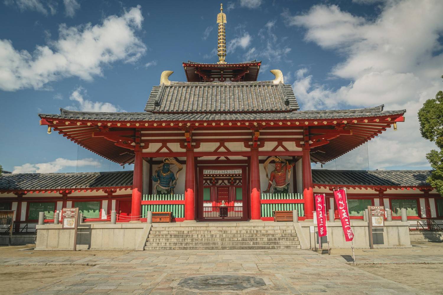 This image features the vibrant red Chumon (Inner Gate) of Shitennō-ji Temple in Osaka, characterized by its traditional Japanese tiered roof and golden finial. Guarding the entrance are two imposing Nio statues, colored blue and red, set against a bright blue sky with scattered clouds.