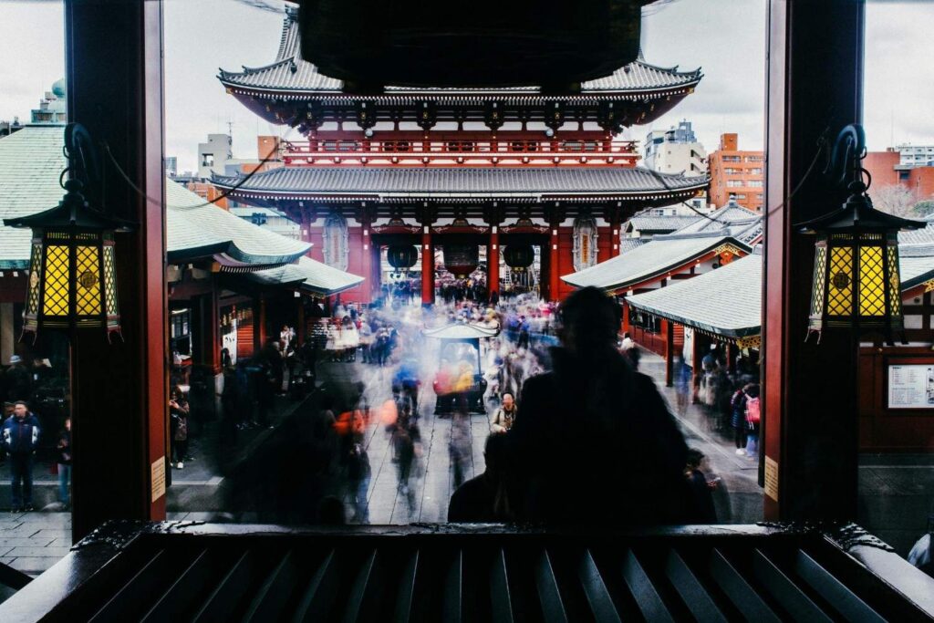 This long-exposure photograph captures the bustling courtyard of Sensō-ji Temple in Tokyo, framed by dark foreground pillars and glowing yellow lanterns. A blurred crowd of visitors flows between the traditional tiered gates and smaller surrounding structures, creating a sense of constant movement within the sacred space.
