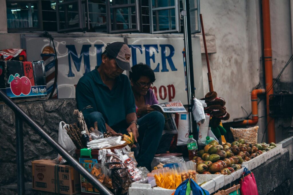 A man and a woman sit together behind a street-side fruit stall, with the man focused on peeling fruit while the woman reads a newspaper. Their small shop is filled with an array of fresh mangoes, packaged snacks, and crates of produce under a weathered building sign.