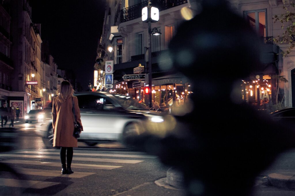 A woman in a tan coat stands at a crosswalk as a car blurs past her on a busy city street at night. The scene captures a vibrant urban atmosphere, with glowing cafe lights and street signs illuminating the dark surroundings.