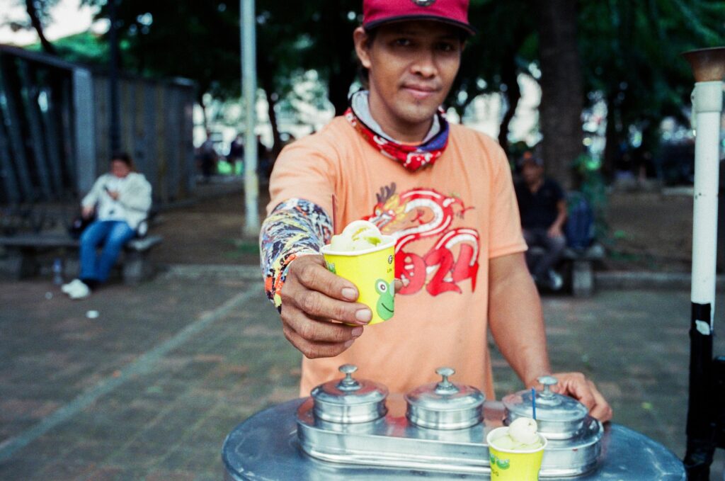 A street vendor wearing a red cap and orange shirt offers a small yellow cup of lime-colored sorbet toward the camera. He stands behind a silver cart with metal containers in a park setting with trees and blurred figures in the background.