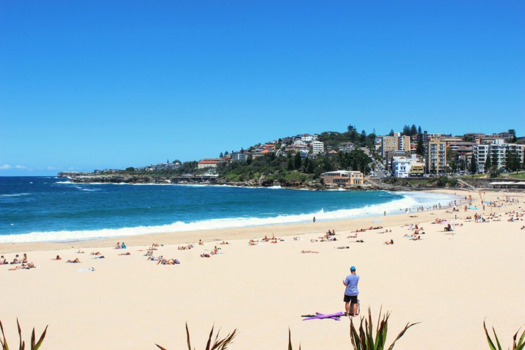 A vibrant, sun-drenched beach is filled with people lounging on golden sand against a backdrop of rolling turquoise waves. In the distance, a lush hillside is densely packed with coastal buildings under a clear, bright blue sky.