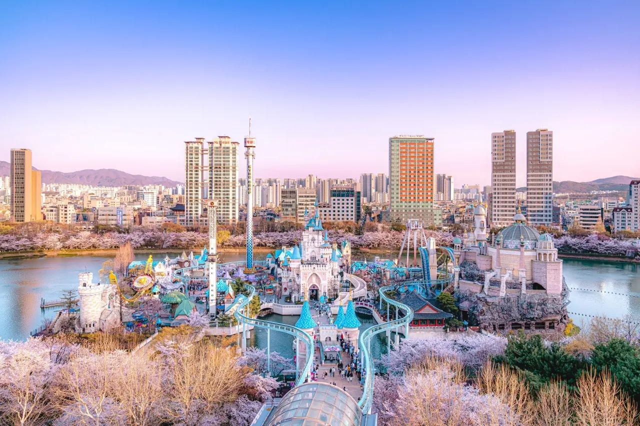 This wide-angle shot captures the colorful Lotte World theme park in Seoul, nestled within a urban landscape surrounded by blooming cherry blossoms and high-rise buildings. The scene features fairytale-inspired castles and amusement rides situated on an island in the middle of a calm lake under a soft, pastel sky.