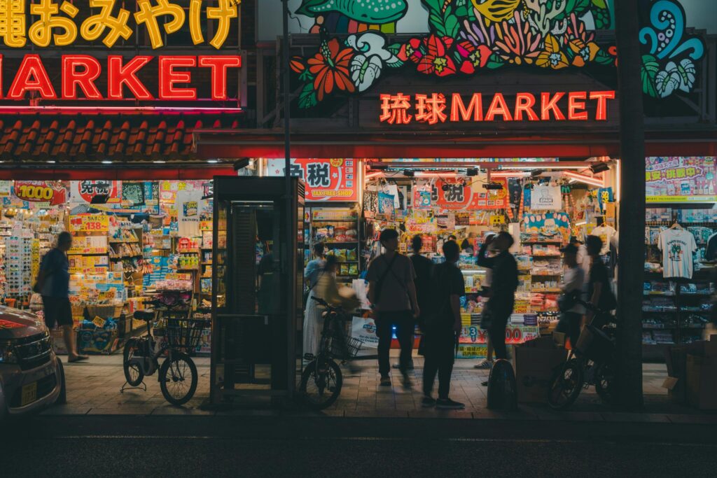 This night scene captures the glowing neon storefronts of "Ryukyu Market," a vibrant Japanese shop packed with colorful souvenirs and household goods. Pedestrians and bicycles are silhouetted against the bright interior lights, creating a bustling atmosphere on the city sidewalk.