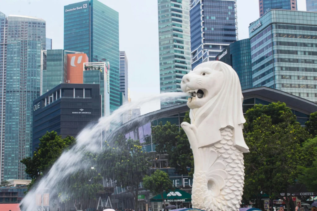 The Merlion statue stands prominently in the foreground, spouting a steady stream of water against the backdrop of Singapore's urban skyline. Modern skyscrapers featuring prominent corporate logos rise behind the iconic monument under a soft, overcast sky.