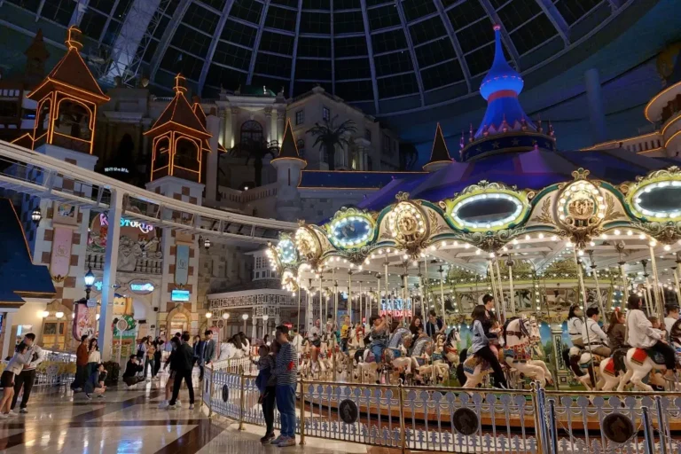 An ornate carousel glows with warm lights under a massive glass dome in a bustling indoor theme park. Visitors stroll through the whimsical, castle-like environment as a white monorail track winds overhead.