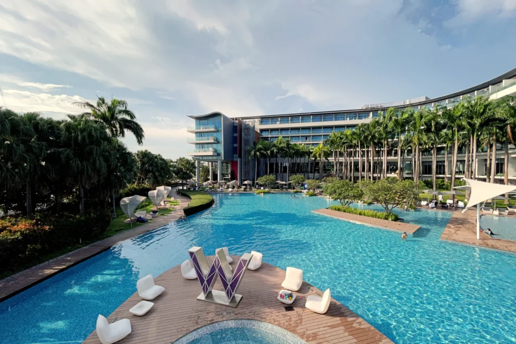 This image shows a large, winding swimming pool at a luxury resort, featuring a prominent "W" sculpture on a wooden deck in the foreground. The scene is framed by modern hotel architecture and lush palm trees under a bright, partly cloudy sky.