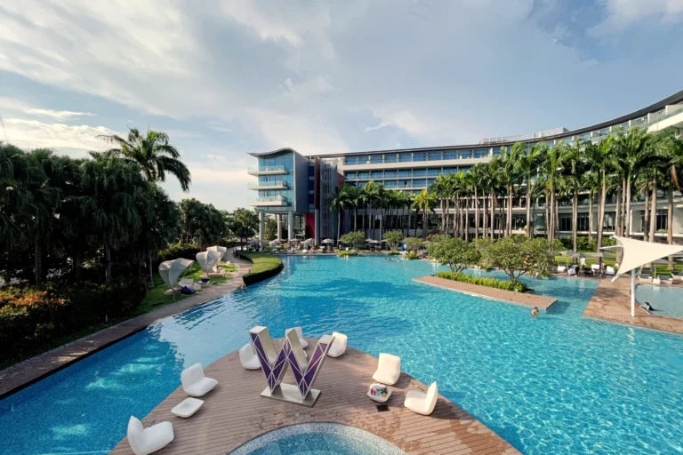This image shows a large, winding swimming pool at a luxury resort, featuring a prominent "W" sculpture on a wooden deck in the foreground. The scene is framed by modern hotel architecture and lush palm trees under a bright, partly cloudy sky.