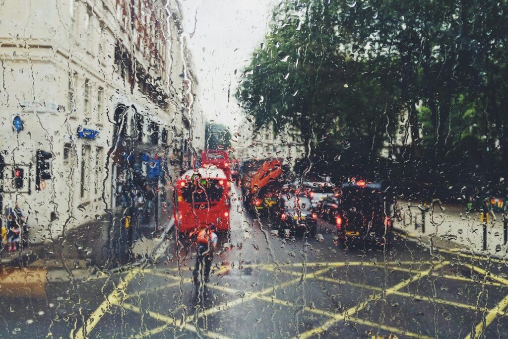This image captures a rain-streaked view of a busy London street through a window, with iconic red double-decker buses and cars blurred in the traffic. The heavy droplets and running water on the glass create a textured, atmospheric filter over the urban scene and the green trees lining the road.