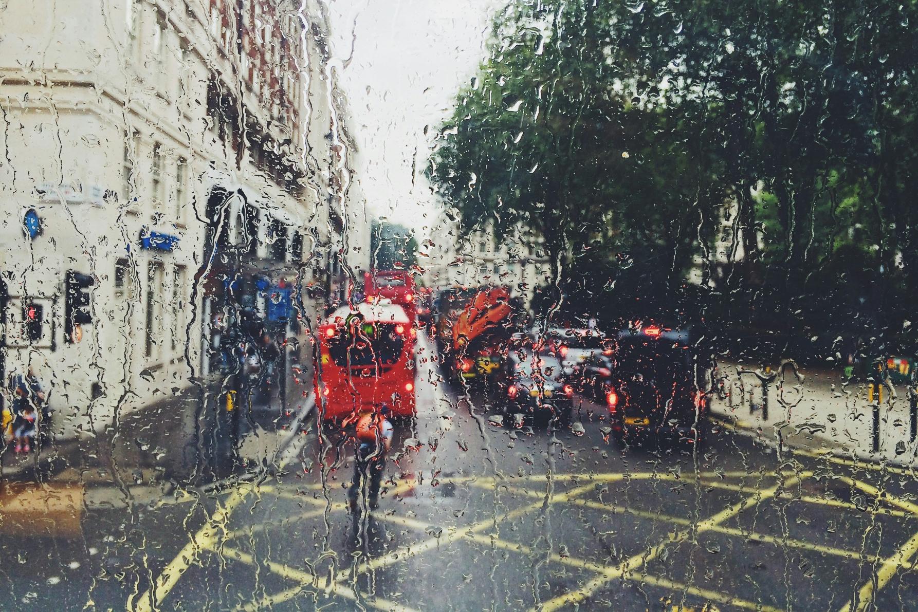 This image captures a rain-streaked view of a busy London street through a window, with iconic red double-decker buses and cars blurred in the traffic. The heavy droplets and running water on the glass create a textured, atmospheric filter over the urban scene and the green trees lining the road.