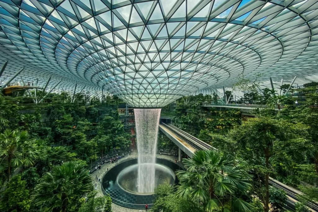 A massive indoor waterfall cascades from the center of a geometric glass dome into a lush, terraced forest. A modern train track curves through the vibrant greenery surrounding the central water feature.