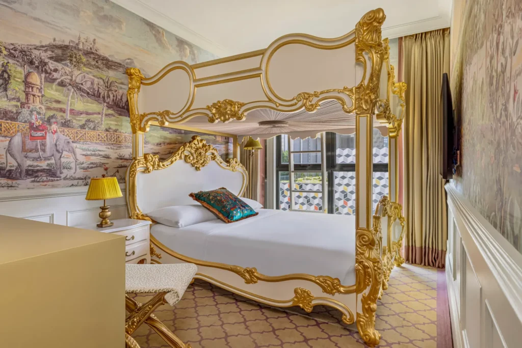 This ornate bedroom features a lavish white and gold rococo-style canopy bed positioned against a large, detailed mural of an elephant and a tropical landscape. The space is accented by patterned carpeting, a vintage-style yellow lamp, and wainscoted walls that create a highly decorative, regal atmosphere.