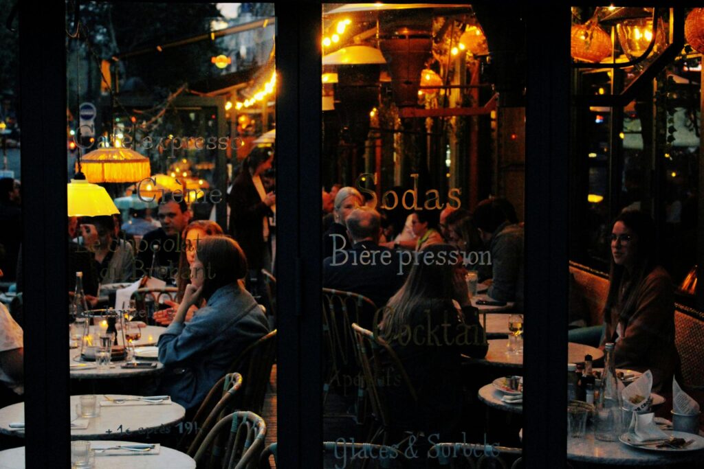 This image captures a warm, dimly lit evening scene at a French café, viewed through glass windows etched with gold lettering for drinks and snacks. Patrons are gathered at small round tables under glowing amber lamps, creating a cozy and bustling atmosphere typical of a European terrace.