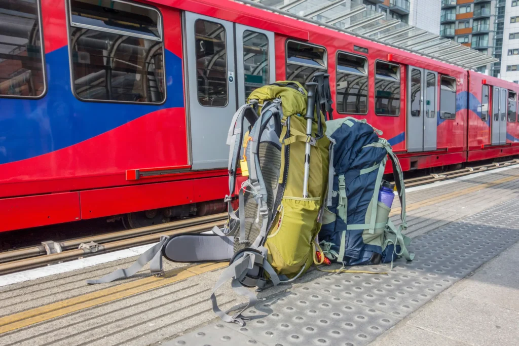 Two large hiking backpacks and trekking poles sit on a train platform next to a modern red and blue commuter train. The outdoor station features tactile paving and glass-roofed shelters, suggesting the beginning or end of a long-distance journey.
