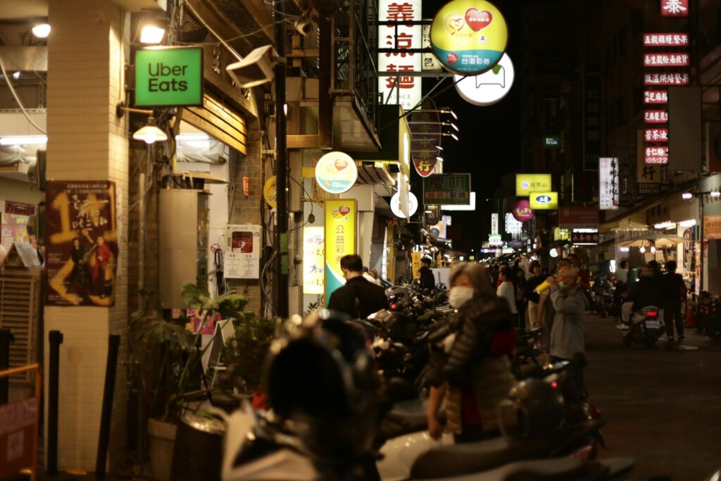 A bustling night scene captures a narrow street in Taiwan lined with vibrant glowing signs and rows of parked scooters. People wearing masks walk past various storefronts, including one marked with a bright green Uber Eats sign.