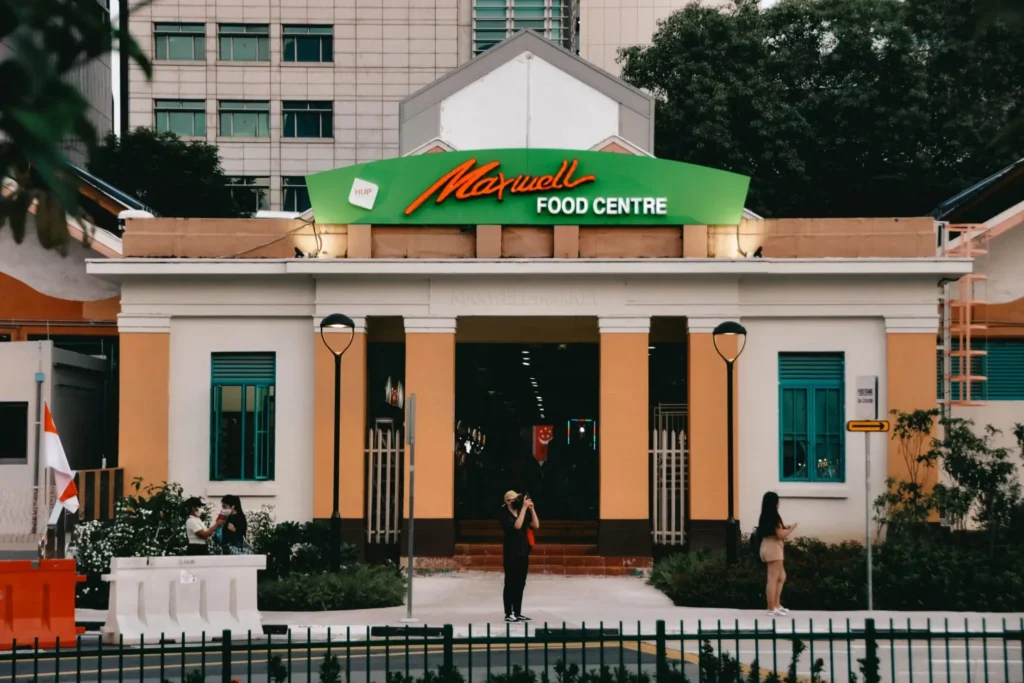 The image shows the green and orange exterior of the Maxwell Food Centre in Singapore. Several people are standing on the sidewalk in front of the building’s entrance.
