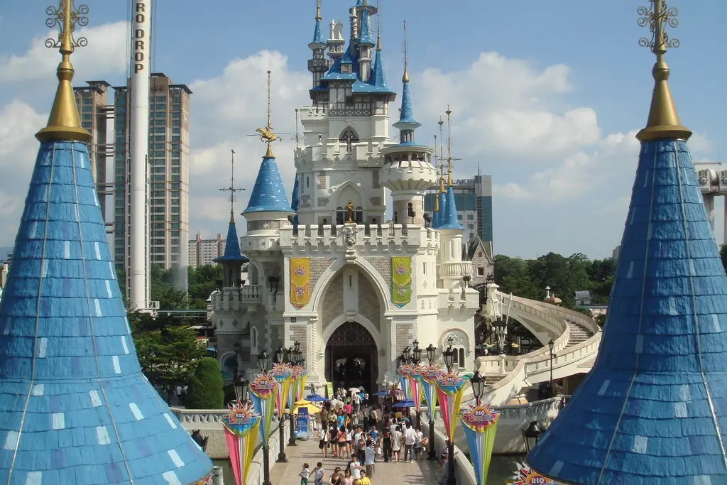 A fairy-tale castle with white stone walls and bright blue spires stands as the centerpiece of an outdoor theme park. Crowds of people walk across a bridge toward the arched entrance, framed by colorful banners and set against a backdrop of modern high-rise buildings.
