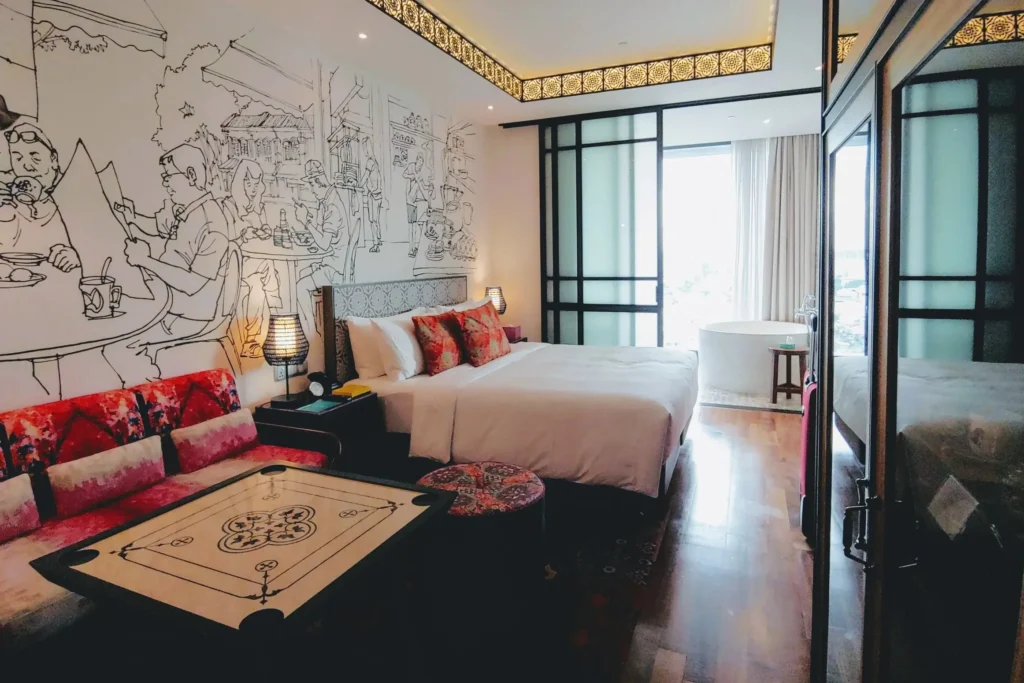 This modern hotel room features a large illustrated wall mural depicting a street cafe scene alongside a bed with vibrant red patterned pillows. The space includes unique furniture like a carrom board table and an open bathroom area with a standalone circular tub.