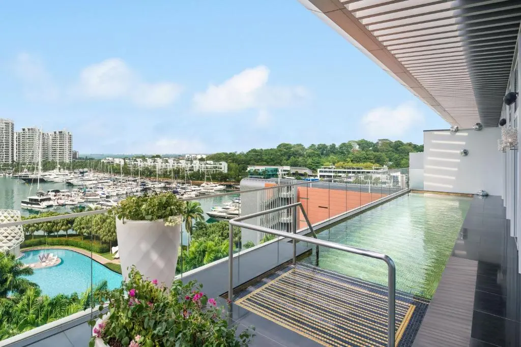 A sleek rooftop terrace features a narrow infinity pool overlooking a vibrant marina filled with yachts and coastal greenery. Modern architecture and lush potted plants frame the view, which extends toward high-rise buildings under a clear, bright sky.