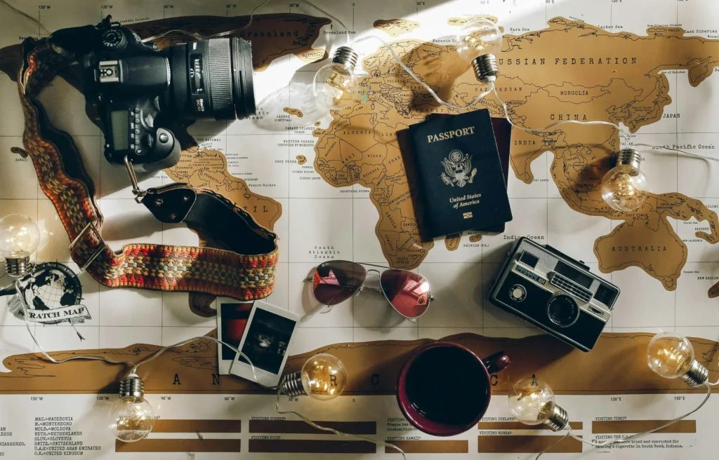 A flat lay captures various travel essentials, including cameras, a passport, and sunglasses, spread across a golden world map illuminated by string lights. The scene is completed by a cup of coffee and small instant photos, evoking a nostalgic and adventurous atmosphere for trip planning.