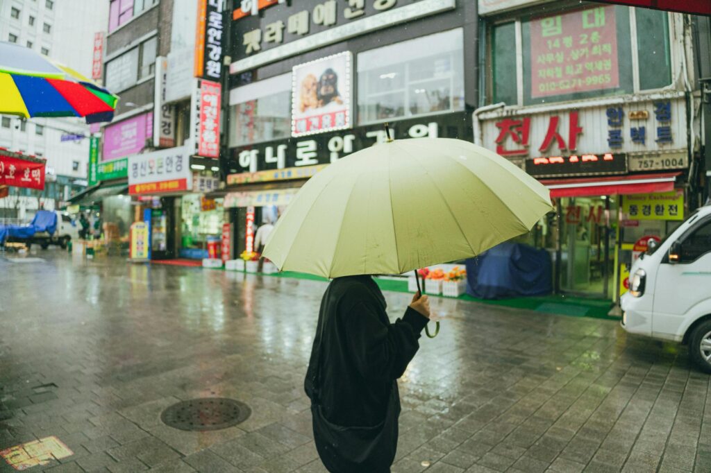 A person in dark clothing stands on a rain-slicked city street, their face hidden beneath a large, pale yellow umbrella. The background features a vibrant array of Korean storefront signs and wet cobblestones that reflect the overcast daylight.