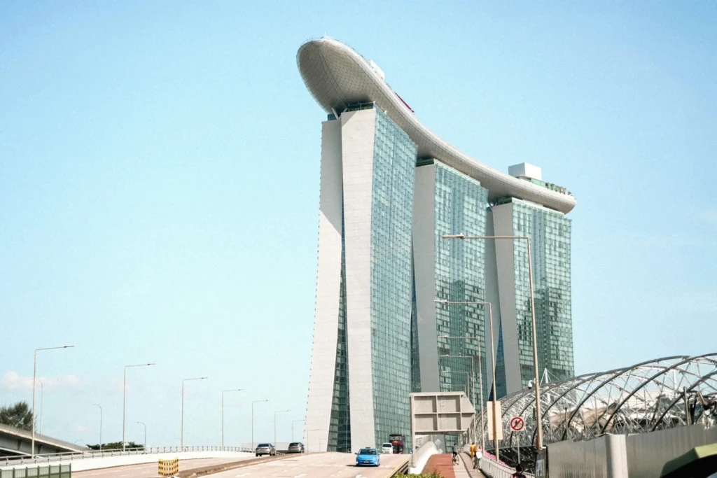 The Marina Bay Sands hotel stands tall against a clear blue sky in Singapore. A wide road with several cars leads toward the iconic, ship-like structure.