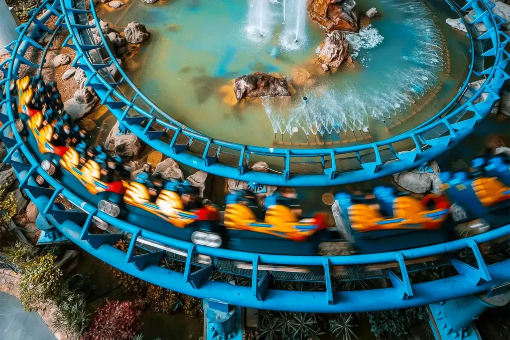 A yellow roller coaster train speeds along a vibrant blue track, creating a dynamic motion blur as it curves around a rocky fountain. The aerial perspective captures the contrast between the high-energy ride and the tranquil, turquoise water feature below.