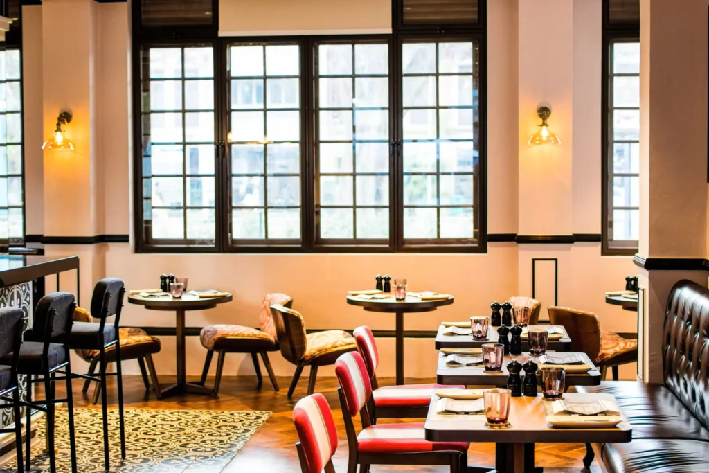 This image showcases the interior of a stylish restaurant featuring warm lighting, large window panes, and eclectic seating options. The design blends modern elements with traditional patterns, seen in the intricate floor tiles and colorful upholstery.