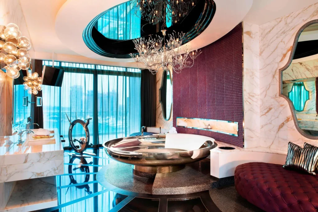 This luxurious room features a striking metallic circular bathtub positioned beneath an ornate crystal chandelier and a reflective ceiling. Marble walls, textured purple accents, and large windows overlooking a cityscape create a sophisticated and modern atmosphere.