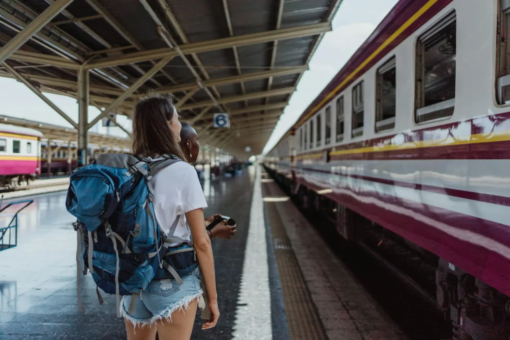 A woman with a large blue hiking backpack stands on a train platform alongside her companion as they wait for their departure. The scene captures a moment of anticipation, with the long, maroon-striped train carriages stretching into the distance under the station's industrial roof.