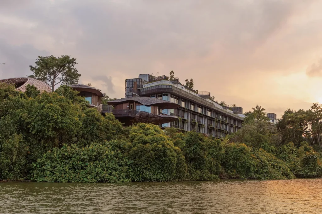 A modern architectural resort featuring organic, seed-pod-shaped structures sits nestled within a lush rainforest canopy along the water's edge. The scene is captured at dusk, with soft orange light glowing behind the biophilic buildings and reflecting off the calm river surface.