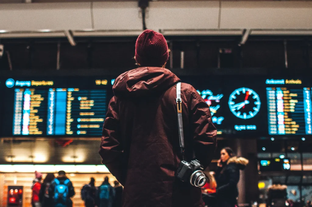 A lone traveler in a maroon beanie and parka stands before a glowing flight information board at a bustling transit hub. With a professional camera slung over one shoulder, they appear to be scanning the departures while other passengers move through the blurred background.