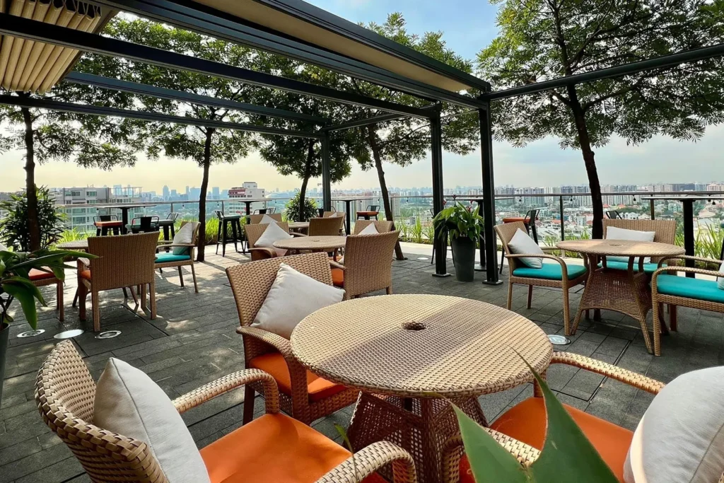 This image showcases a vibrant rooftop terrace with several round wicker tables and chairs featuring bright orange and turquoise cushions. Lush green trees and a modern black pergola frame a wide panoramic view of a city skyline in the distance.
