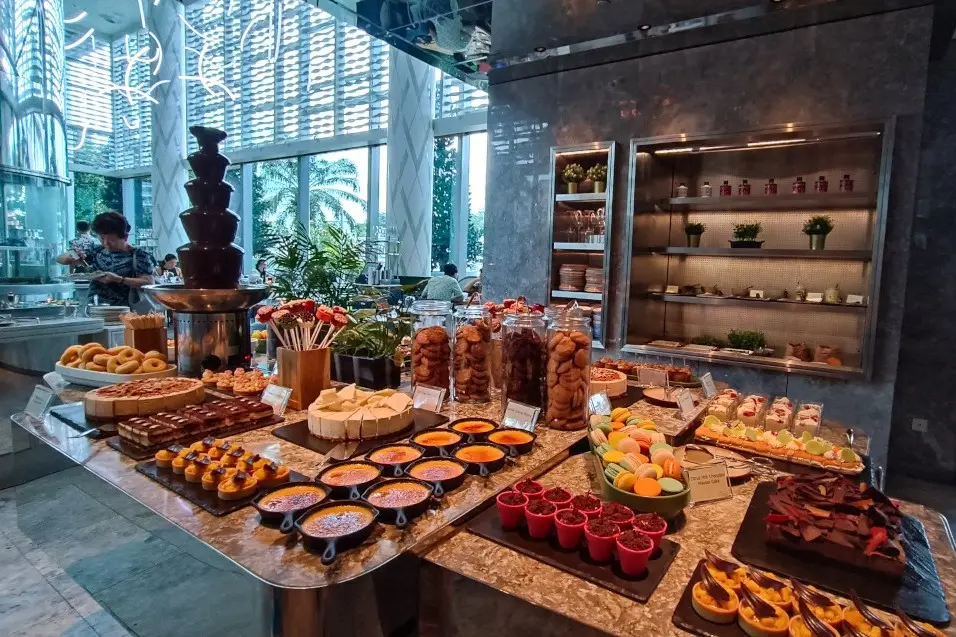 This image showcases an extensive dessert buffet featuring a chocolate fountain, various cakes, macarons, and individual servings of crème brûlée. The spread is elegantly arranged on marble countertops within a modern, sunlit dining area with high ceilings and large windows.