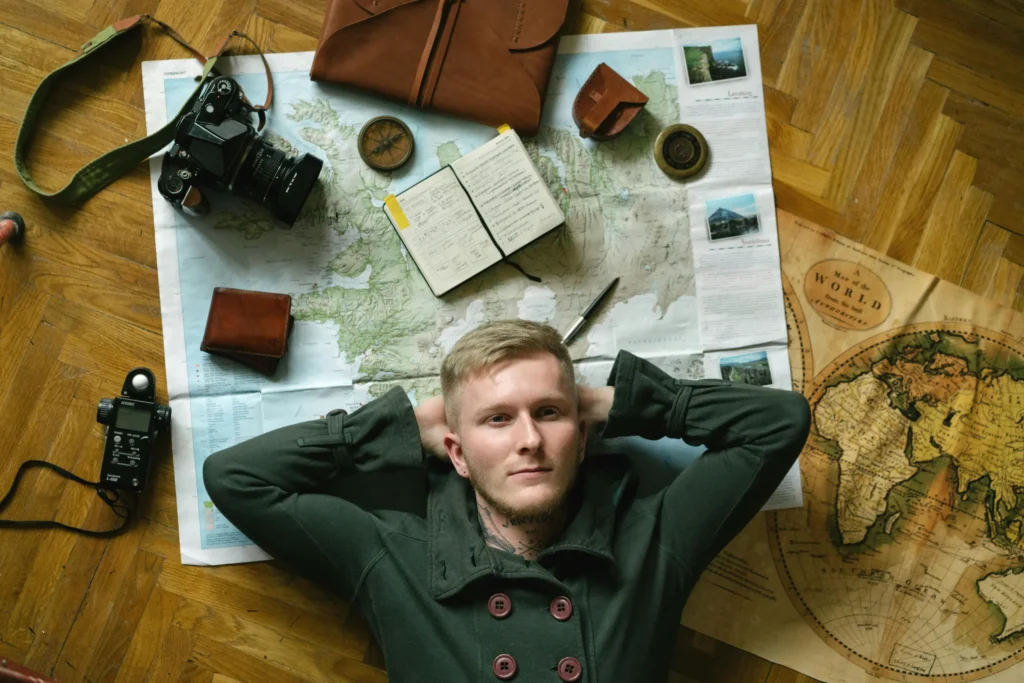 A young man lies on a wooden floor with his hands behind his head, surrounded by travel essentials like maps, a vintage camera, and a journal. The top-down composition and warm lighting create a reflective, adventurous atmosphere focused on trip planning.